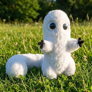 May include: A white plush toy creature with large, round black eyes and small black hands and feet. The toy has a long, curved tail and is set in a grassy field with a blurred background of trees.