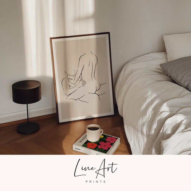 Puede incluir: Un grabado enmarcado de arte lineal que representa dos figuras abraz&aacute;ndose. La obra de arte minimalista utiliza l&iacute;neas negras sobre un fondo beige. Una l&aacute;mpara negra, una taza de caf&eacute; y un libro con im&aacute;genes florales est&aacute;n en primer plano. El texto "Line Art Prints" est&aacute; en la parte inferior.