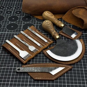 May include: A collection of leatherworking tools, including various stitching chisels, a half-moon knife, and a skiving knife. The tools have wooden handles and are displayed on a black cutting mat with a brown leather background.