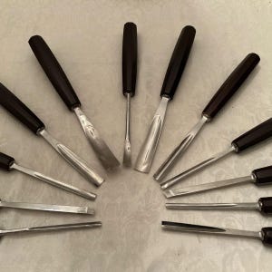 May include: A selection of woodworking chisels arranged in a semi-circle. Each chisel has a polished steel blade and a dark brown, rounded handle. The blades vary in width and shape, indicating a set for detailed carving and shaping of wood.