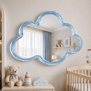 Puede incluir: Un espejo azul claro en forma de nube cuelga de una pared en una guardería. El marco del espejo tiene un diseño ondulado y texturizado. La habitación está decorada con animales de peluche, una cuna y un móvil.