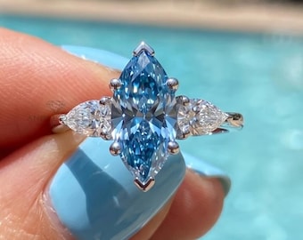 May include: A marquise-cut, light blue gemstone ring with two pear-shaped white gemstones on each side. The ring is held up against a blue background, with the words "ANGLE DUST" visible.