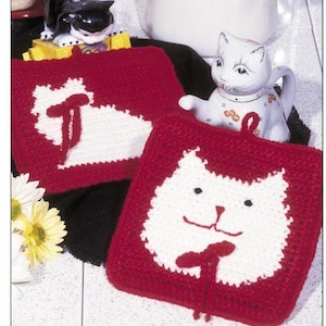 May include: Two red and white cat-themed potholders with a knitted design. The potholders feature a white cat face on a red background. A white ceramic cat figurine is also visible.