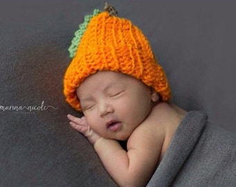 May include: A newborn baby wearing a crocheted orange pumpkin hat with green leaves. The baby is sleeping on a gray blanket.