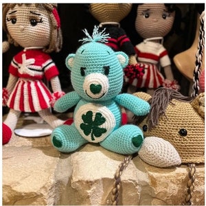 May include: A crocheted blue Care Bear with a green four-leaf clover on its chest. The bear is sitting on a stone surface with other crocheted toys.