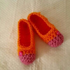 Könnte beinhalten: Ein Paar gehäkelte Hausschuhe in Orange und Rosa mit einem Knopf an jeder Zehe.