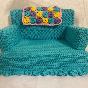 May include: A teal crocheted armchair with a pink, yellow, and purple crocheted throw pillow. The armchair has a ruffled trim around the bottom.
