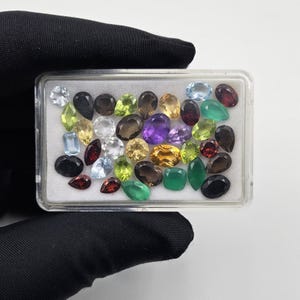 May include: A rectangular clear plastic display case containing a collection of assorted loose faceted gemstones in various shapes, including oval, pear, round, and marquise cuts, featuring colors such as purple, green, yellow, red, and black.
