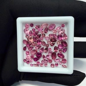May include: A collection of pink gemstones in various shapes and sizes, including oval, pear, and round cuts, are displayed in a white square tray. The gemstones range in color from light pink to deep magenta, with a polished, reflective surface.