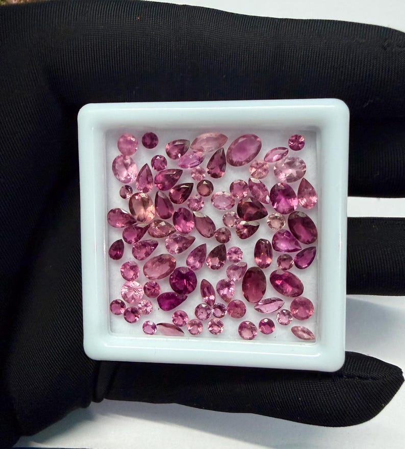May include: A collection of pink gemstones in various shapes and sizes, including oval, pear, and round cuts, are displayed in a white square tray. The gemstones range in color from light pink to deep magenta, with a polished, reflective surface.