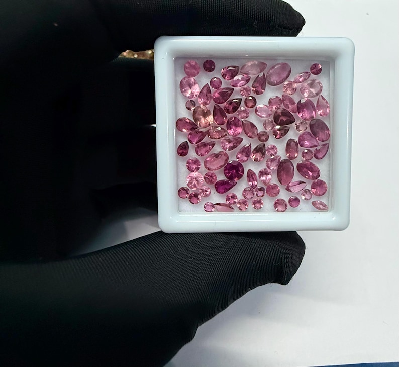 May include: A collection of pink gemstones in various shapes and sizes, including oval, pear, and round cuts, are displayed in a white square container. The gemstones range in color from light to deep pink, with some appearing nearly red. The container is held by a gloved hand.
