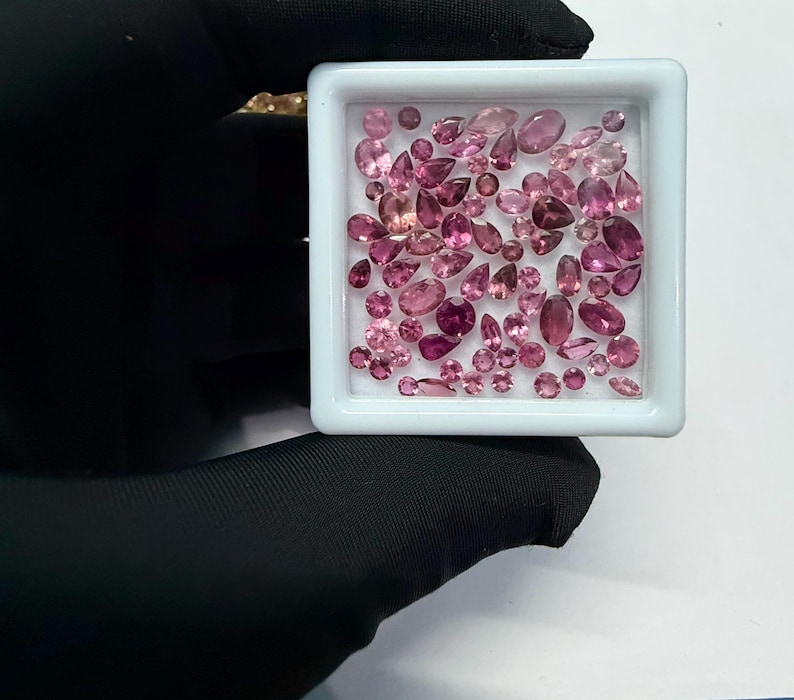 May include: A collection of pink gemstones in various shapes and sizes, including oval, pear, and round cuts, are displayed in a white square tray. The gemstones range in color from light to deep pink. The tray is held by a gloved hand.