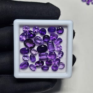 May include: A collection of polished amethyst gemstones in various shapes and sizes, including ovals, rounds, and rectangles, are displayed in a white tray. The gemstones range in color from light to deep purple.