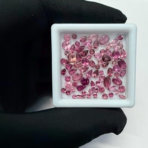 May include: A collection of pink gemstones in various shapes and sizes, including oval, pear, and round cuts, are displayed in a white square container. The gemstones range in color from light to deep pink.
