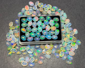 May include: A collection of oval-shaped opals in various colors, including blue, green, and orange. Some opals are contained in a small, rectangular metal box, while others are scattered around it. The opals have a translucent appearance.
