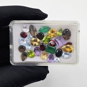 Può includere: Una scatola espositiva rettangolare in plastica trasparente contenente una collezione di pietre preziose sfaccettate multicolori di varie forme, tra cui ovali, a goccia, rotonde e rettangolari, su un inserto in schiuma bianca.