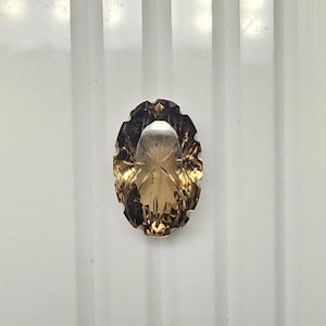 May include: An oval-shaped gemstone with a faceted design. The gem has a warm, golden-brown hue with a lighter centre. The edges are intricately cut, creating a scalloped border. The gemstone is set against a white background.