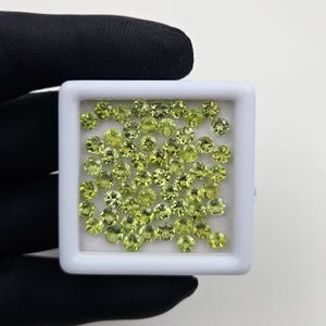 May include: A collection of small, round, light green gemstones displayed in a white square container. The gemstones have a faceted cut, reflecting light. The container is held against a black background.