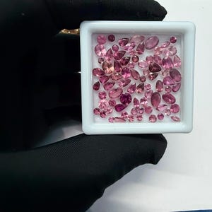 May include: A collection of pink gemstones in various shapes and sizes, including oval, pear, and round cuts, are displayed in a white square container. The gemstones range in color from light to deep pink, with some appearing nearly red. The container is held by a gloved hand.