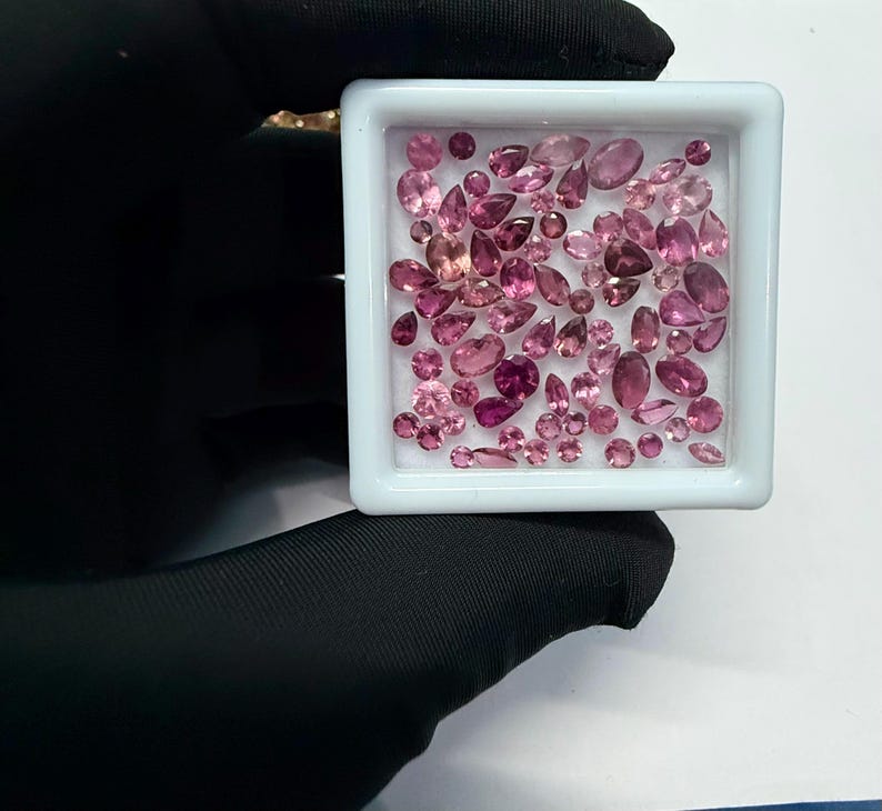 May include: A collection of pink gemstones in various shapes and sizes, including oval, pear, and round cuts, are displayed in a white square container. The gemstones range in color from light to deep pink, with some appearing nearly red. The container is held by a gloved hand.