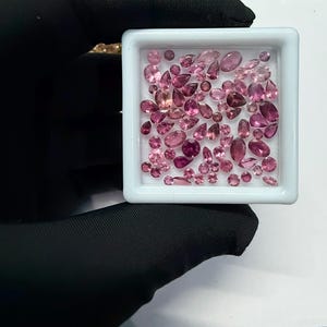 May include: A collection of pink gemstones in various shapes and sizes, including oval, pear, and round cuts, are displayed in a white square tray. The gemstones range in color from light to deep pink. The tray is held by a gloved hand.
