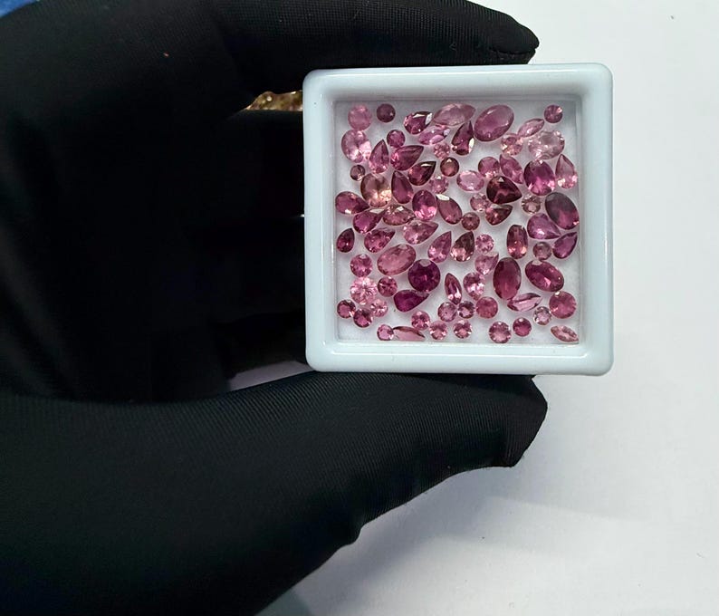 May include: A collection of pink gemstones in various shapes and sizes, including oval, pear, and round cuts, are displayed in a white square tray. The gemstones range in color from light to deep pink. The tray is held by a gloved hand.