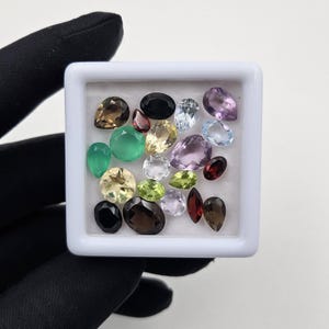May include: A collection of assorted loose faceted gemstones in various shapes and colors, including green, brown, purple, red, yellow, and clear stones, arranged inside a square white display box with a white foam insert for organization.