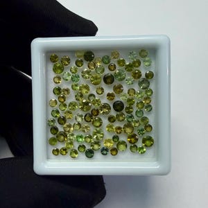 Könnte beinhalten: Eine Sammlung kleiner, runder, facettierter Edelsteine in Grün- und Gelbgrüntönen. Die Edelsteine befinden sich in einer weißen, quadratischen Schale. Die Edelsteine variieren in Größe und Farbe und bilden eine ansprechende Darstellung.