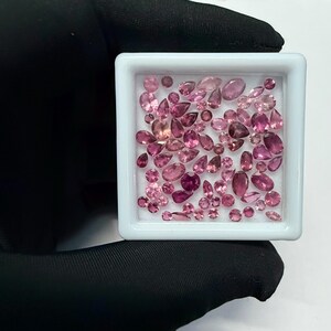May include: A collection of pink gemstones in various shapes and sizes, including oval, pear, and round cuts, are displayed in a white square container. The gemstones range in color from light to deep pink.