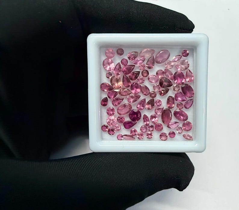 May include: A collection of pink gemstones in various shapes and sizes, including oval, pear, and round cuts, are displayed in a white square container. The gemstones range in color from light to deep pink.