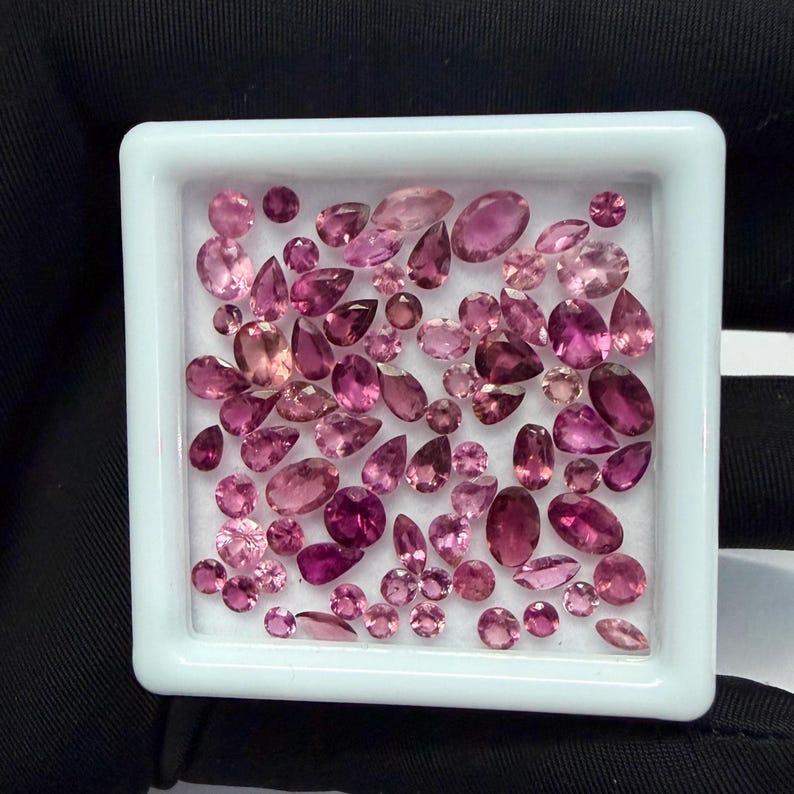 May include: A collection of pink gemstones in various shapes and sizes, including oval, pear, and round cuts, are displayed in a white square tray. The gemstones range in color from light pink to deep magenta, creating a vibrant display.