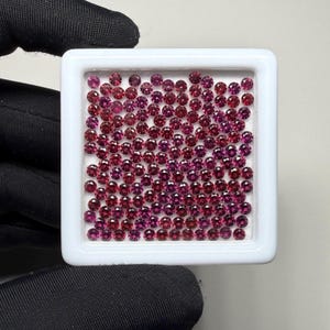 May include: A white square tray filled with numerous small, round gemstones in varying shades of red and purple. The gemstones are closely packed together, creating a dense and colourful display. The image is well-lit, showcasing the gems' facets and brilliance.