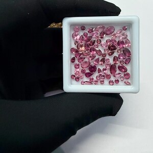 May include: A collection of pink gemstones in various shapes and sizes, including oval, pear, and round cuts, are displayed in a white square tray. The gemstones range in shades from light pink to deep magenta, showcasing a variety of hues and brilliance.