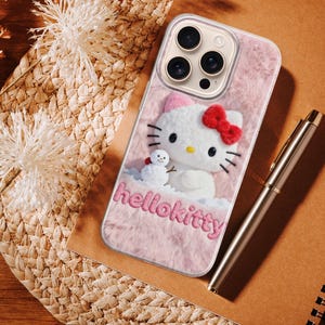 May include: A pink phone case featuring a Hello Kitty design with a red bow and a small snowman. The case has the text "hellokitty" in pink. A silver pen and a spiral notebook are also visible.