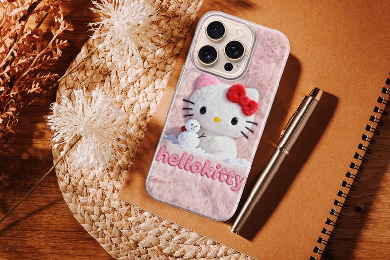 May include: A pink phone case featuring a Hello Kitty design with a red bow and a small snowman. The case has the text "hellokitty" in pink. A silver pen and a spiral notebook are also visible.