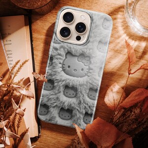 May include: A gray phone case featuring a plush, furry texture and embossed Hello Kitty faces. The case has a camera cutout and is surrounded by a wooden surface, dried leaves, and a glass of water.