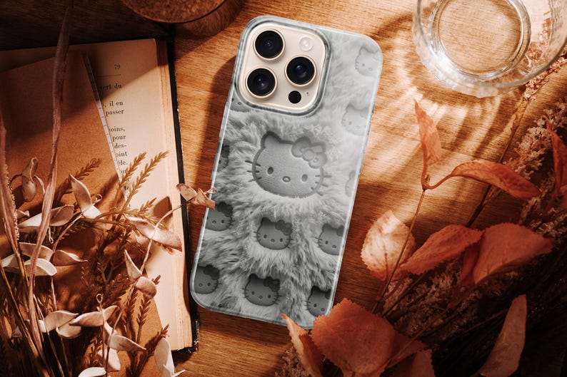 May include: A gray phone case featuring a plush, furry texture and embossed Hello Kitty faces. The case has a camera cutout and is surrounded by a wooden surface, dried leaves, and a glass of water.