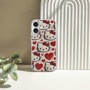 May include: A clear phone case featuring a repeating pattern of Hello Kitty faces with red bows and hearts. The case has a glossy finish and is designed for a smartphone. The background includes a white arched decor and a neutral-toned wall.