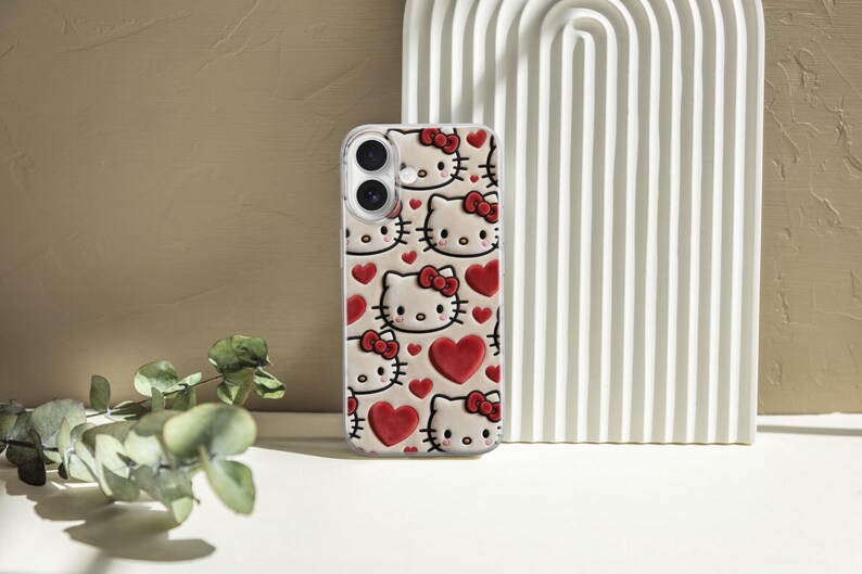 May include: A clear phone case featuring a repeating pattern of Hello Kitty faces with red bows and hearts. The case has a glossy finish and is designed for a smartphone. The background includes a white arched decor and a neutral-toned wall.