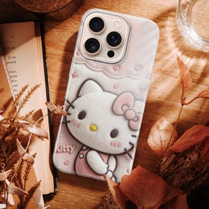 May include: A pink phone case featuring a 3D image of Hello Kitty with a bow, hearts, and the word "Kitty". The case is on a wooden surface with a book, dried leaves, and a glass of water.