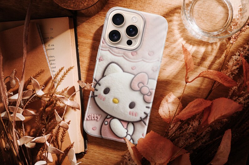 May include: A pink phone case featuring a 3D image of Hello Kitty with a bow, hearts, and the word "Kitty". The case is on a wooden surface with a book, dried leaves, and a glass of water.