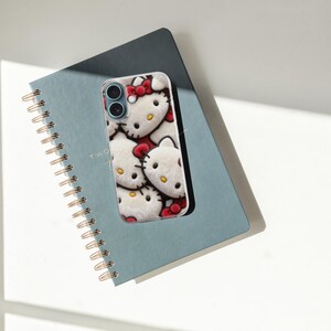 May include: A white and red Hello Kitty phone case rests on a blue spiral notebook. The phone case features a repeating pattern of Hello Kitty's face with a red bow. The notebook has gold spiral binding and the word "TWO" printed on the cover.