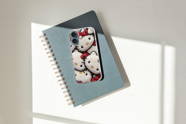 May include: A white and red Hello Kitty phone case rests on a blue spiral notebook. The phone case features a repeating pattern of Hello Kitty's face with a red bow. The notebook has gold spiral binding and the word "TWO" printed on the cover.