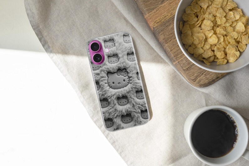 May include: A phone case featuring a repeating pattern of Hello Kitty faces on a gray, fluffy background. The case has a pink camera lens surround. The image also shows a bowl of cereal, a cup of coffee, and a wooden cutting board.