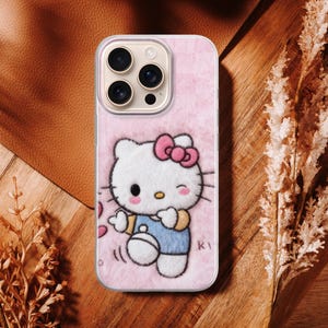 May include: Pink phone case featuring a Hello Kitty design. The case has a soft, fuzzy texture and a cartoon illustration of Hello Kitty wearing a blue outfit and a pink bow. The case has a camera cutout and is surrounded by a brown and wooden background.