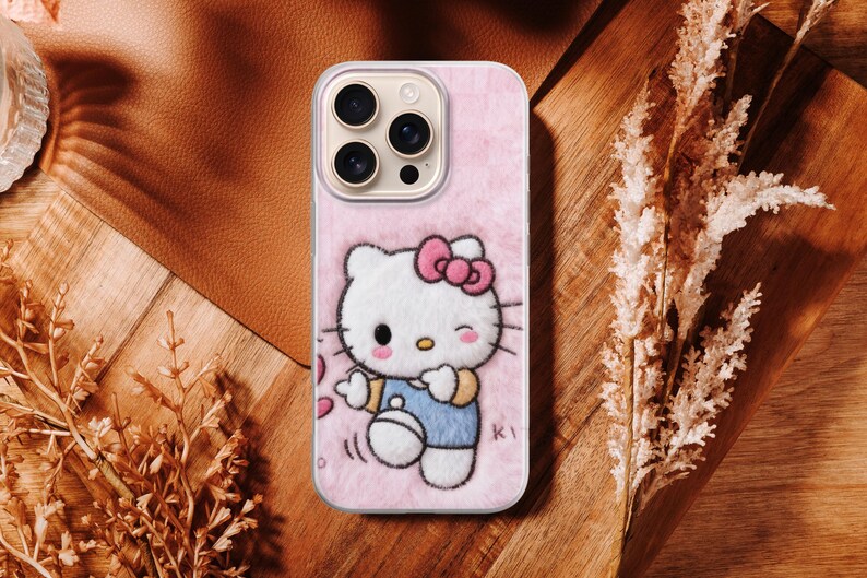 May include: Pink phone case featuring a Hello Kitty design. The case has a soft, fuzzy texture and a cartoon illustration of Hello Kitty wearing a blue outfit and a pink bow. The case has a camera cutout and is surrounded by a brown and wooden background.