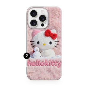 May include: A pink, fluffy phone case featuring a Hello Kitty design. The case has a white Hello Kitty character with a red bow and a small snowman. The text "hellokitty" is embroidered at the bottom.
