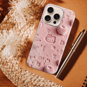 May include: A pink phone case featuring a Hello Kitty design with embossed apples and stars. The case is on a brown surface next to a silver pen and a spiral notebook. Decorative dried flowers and woven straw mat are in the background.