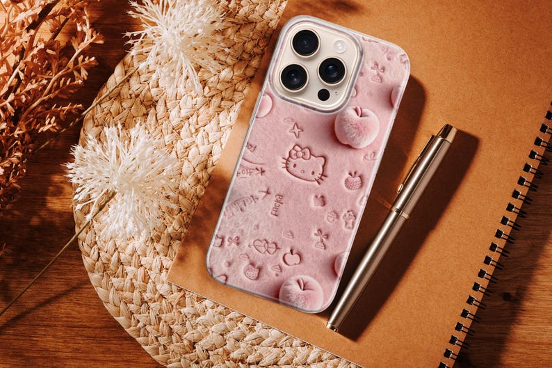May include: A pink phone case featuring a Hello Kitty design with embossed apples and stars. The case is on a brown surface next to a silver pen and a spiral notebook. Decorative dried flowers and woven straw mat are in the background.
