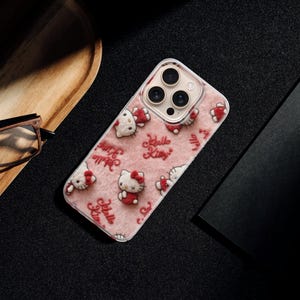 May include: A pink phone case featuring a repeating pattern of Hello Kitty characters and the text "Hello Kitty." The case is on a black surface, next to a wooden tray, glasses, and a black notebook.